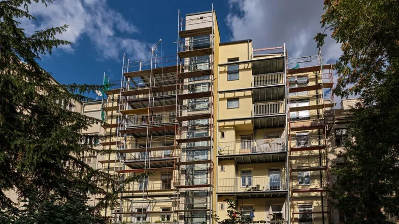 Mehrstöckiges Wohngebäude mit Gerüst an der Fassade, umgeben von Bäumen unter blauem Himmel.