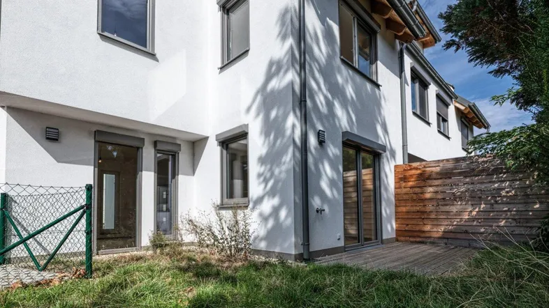 Moderne Hausfassade mit weißem Putz, großen Fenstern und einer Holzterrasse im Gartenbereich.