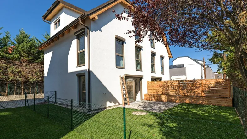 Modernes Einfamilienhaus mit weißer Fassade, Garten und gepflasterter Terrasse im Sonnenlicht.