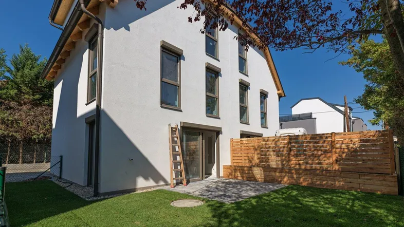 Modernes Einfamilienhaus mit weißer Fassade, gepflasterter Terrasse und grünem Gartenbereich.