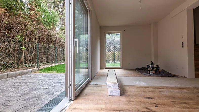 Heller Wohnbereich im Bau mit großen Schiebetüren, die zu einer gepflasterten Terrasse und einem Garten führen.