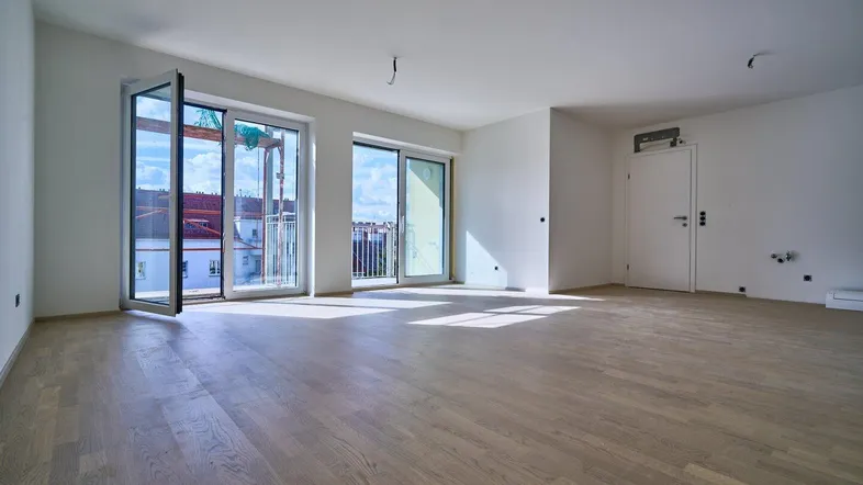 Helles, leeres Zimmer mit Holzboden und großen Fenstern, die Zugang zu einem Balkon bieten.