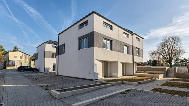 Moderne Reihenhäuser mit weiß-grauer Fassade und gepflasterten Parkflächen unter blauem Himmel.