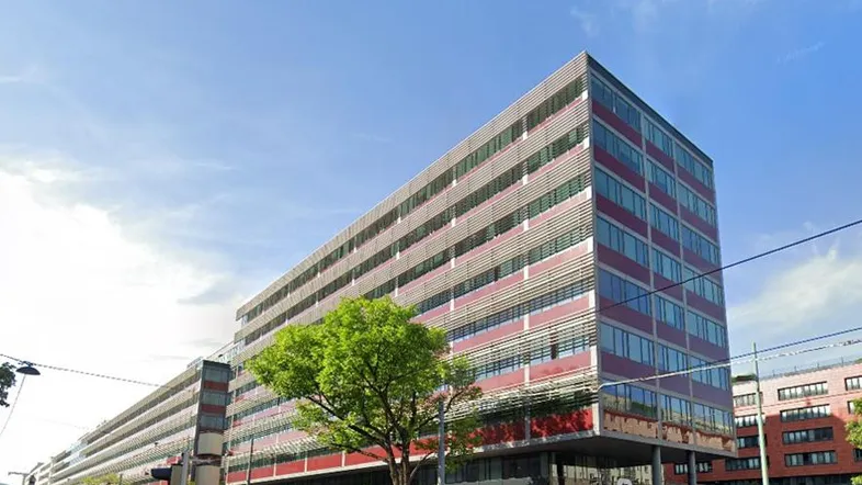 Modernes Bürogebäude mit roter und grauer Fassade unter blauem Himmel in einer städtischen Umgebung.