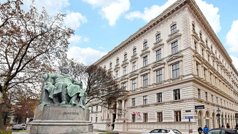 Historisches Gebäude mit klassischer Architektur und einer Statue auf dem Vorplatz unter blauem Himmel.