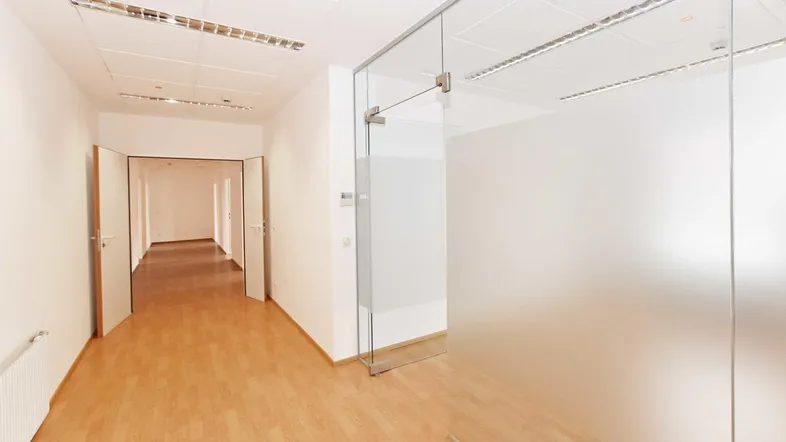 Heller Bürogang mit modernen Glaswänden und hellem Laminatboden, der zu weiteren Büros führt.