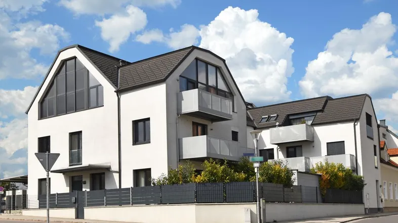 Modernes Mehrfamilienhaus mit weißer Fassade, dunklem Dach und mehreren Balkonen an einer Straßenecke.