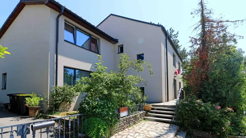 Mehrfamilienhaus mit heller Fassade, umgeben von Bäumen und Sträuchern, mit einem gepflasterten Zugangsweg.