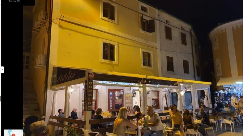 Nachtaufnahme eines Gebäudes mit einem belebten Restaurant im Erdgeschoss und Gästen im Außenbereich.