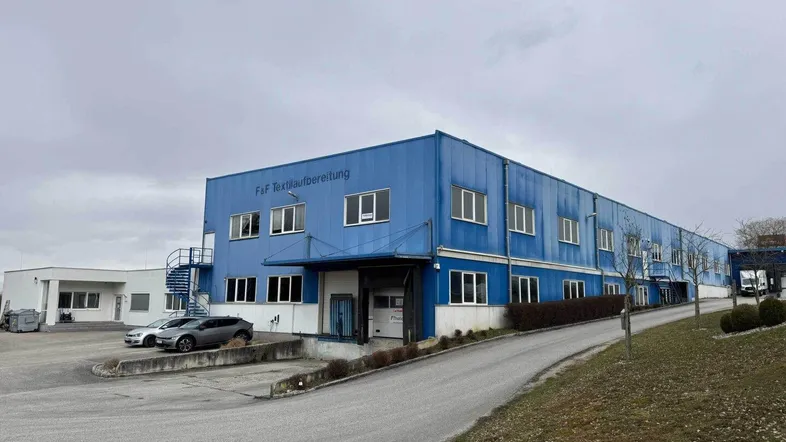 Blaues Industriegebäude mit mehreren Fenstern und einer Zufahrtsrampe unter bewölktem Himmel.