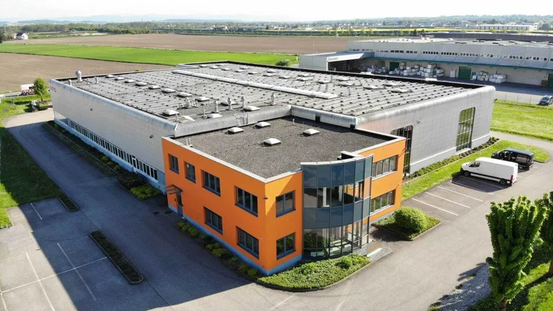 Luftaufnahme eines modernen Industriegebäudes mit orangefarbenem Bürotrakt und weitläufigen Parkplätzen.