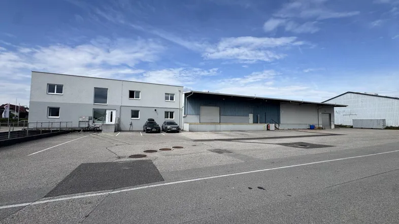 Ansicht des modernen Bürogebäudes und der angrenzenden Lagerhalle mit großem Parkplatz unter blauem Himmel.