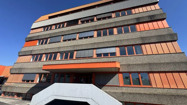 Modernes Bürogebäude mit markanter Betonfassade und orangefarbenen Akzenten unter blauem Himmel.