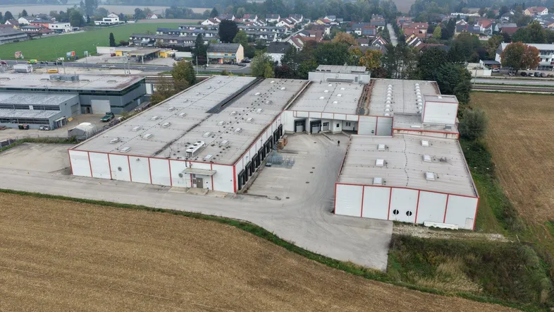 Luftaufnahme eines großen Industriegebäudes mit mehreren Hallen und umliegenden Feldern in ländlicher Umgebung.