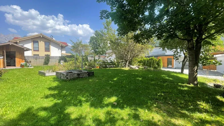 Großer, sonniger Gartenbereich mit Rasenfläche, Bäumen und einem Hochbeet im Hintergrund.
