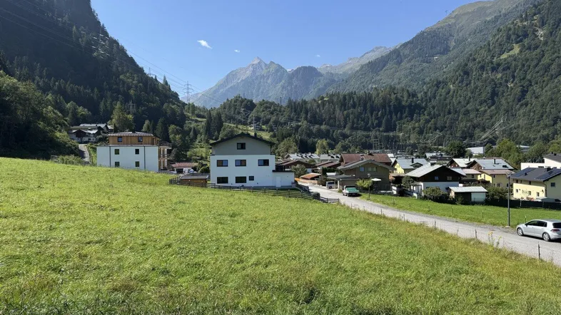 Blick auf ein malerisches Dorf in einem grünen Tal, umgeben von majestätischen Bergen unter blauem Himmel.