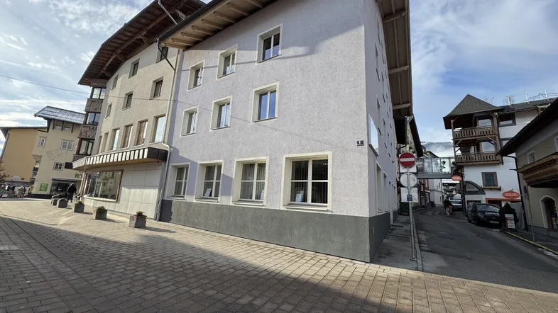 Mehrstöckiges Gebäude mit heller Fassade und dunklem Sockel an einer gepflasterten Straße in einer Bergregion.