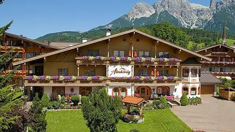 Traditionelles Alpenhotel mit Holzfassade, blühenden Balkonen und gepflegtem Garten vor beeindruckender Bergkulisse.
