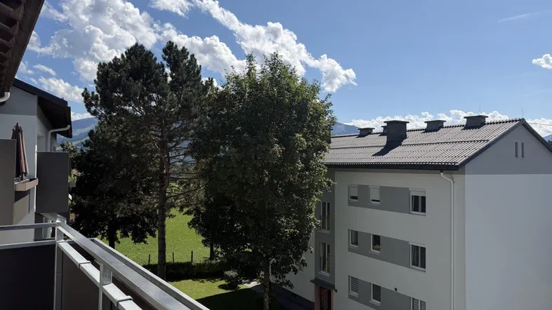 Blick vom Balkon auf grüne Wiesen, Bäume und umliegende Gebäude unter blauem Himmel.