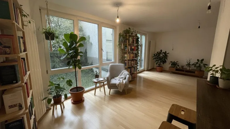 Helles Wohnzimmer mit großen Fenstern, hellem Holzboden, Bücherregalen und vielen Pflanzen.