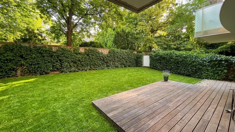 Gepflegter Garten mit Rasenfläche und Holzterrasse, umgeben von üppigem Grün und Hecken.