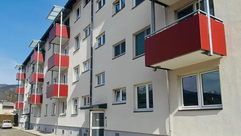 Mehrfamilienhaus mit weißer Fassade und roten Balkonen unter blauem Himmel.