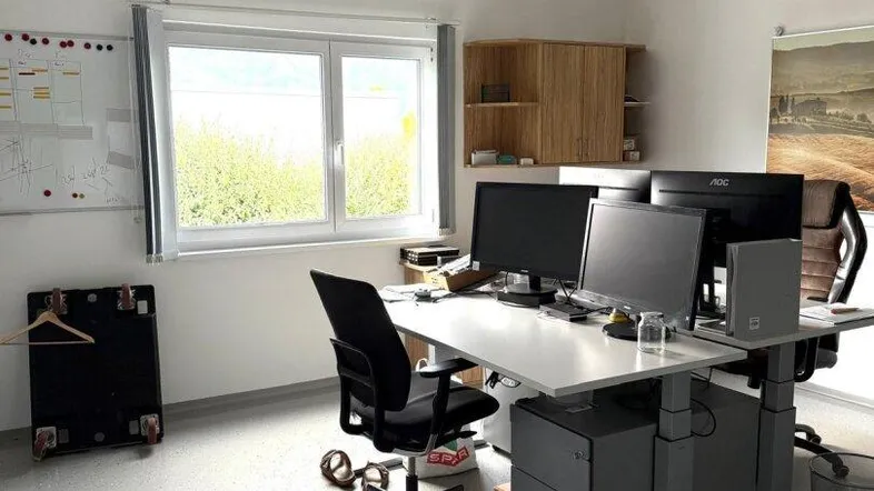 Helles Büro mit modernem Arbeitsplatz, mehreren Monitoren und Blick ins Grüne durch das Fenster.
