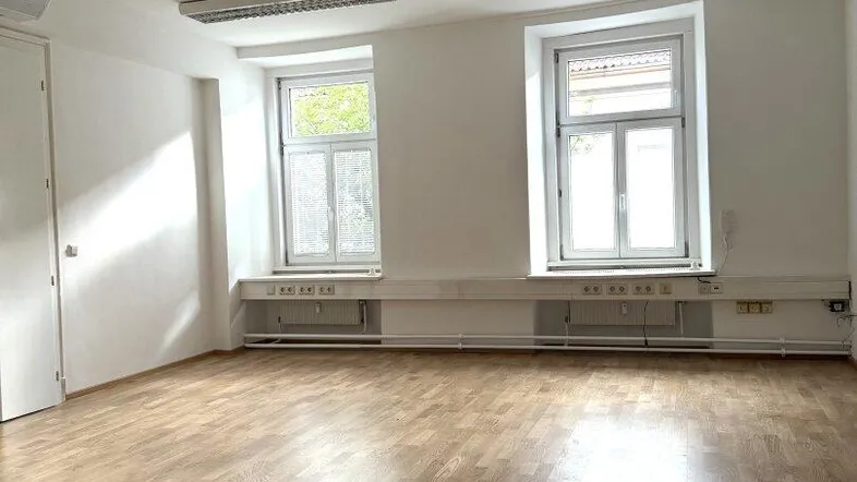 Heller Büroraum mit Holzparkett, weißen Wänden, zwei Fenstern und Klimaanlage.