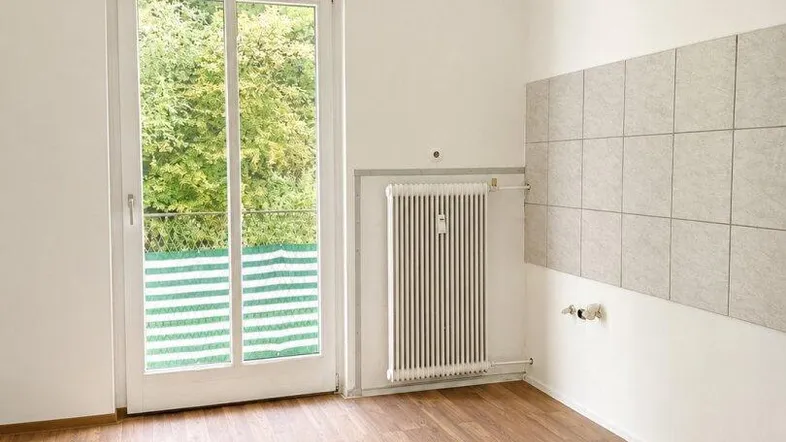 Leerer Küchenraum mit Holzboden, Fliesenwand und Zugang zum Balkon mit Blick ins Grüne.