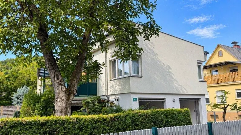 Modernes Haus mit weißem Lattenzaun und altem Baum im Vorgarten unter blauem Himmel.