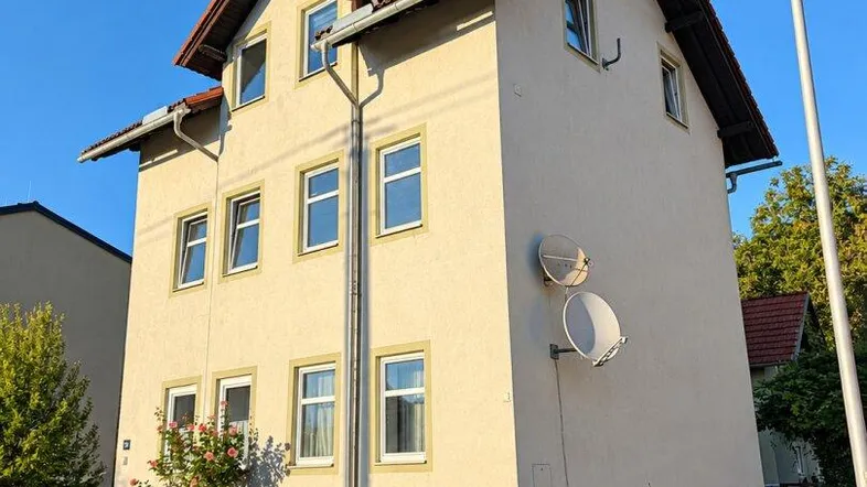 Mehrfamilienhaus mit heller Fassade, Satteldach und Satellitenschüsseln an einem sonnigen Tag.