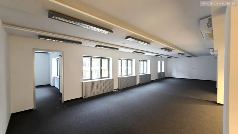 Geräumiger, leerer Büroraum mit Teppichboden, vielen Fenstern und moderner Deckenbeleuchtung.
