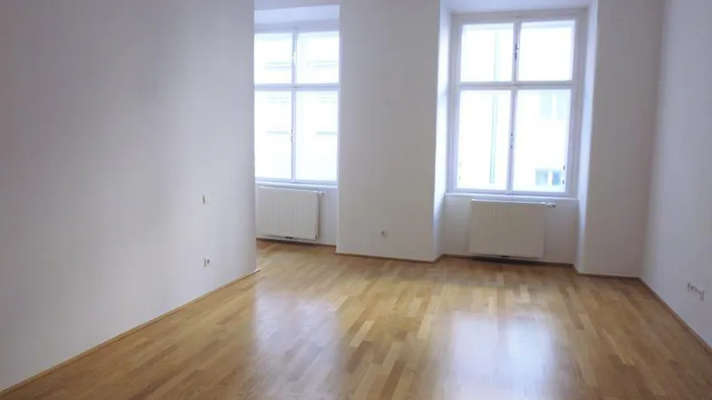 Heller, leerer Raum mit Holzboden und großen Fenstern, der viel Tageslicht hereinlässt.