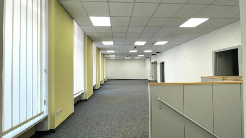 Geräumiges, leeres Büro mit Teppichboden, Rasterdecke und vertikalen Jalousien an den Fenstern.