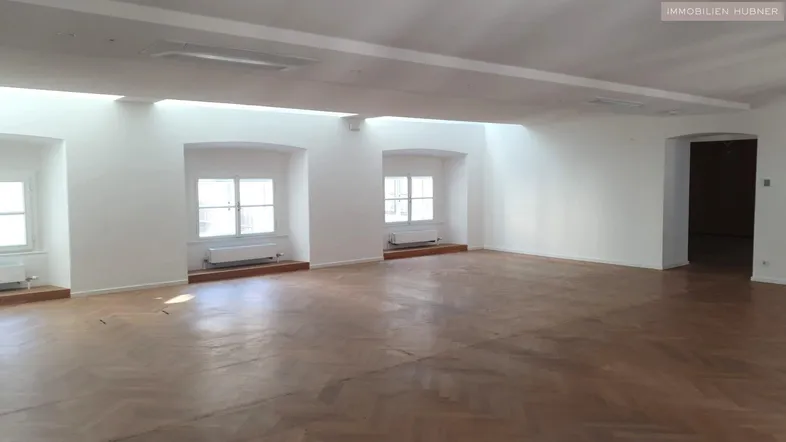 Großer, leerer Raum mit Fischgrätparkettboden und mehreren Fenstern, die viel Tageslicht hereinlassen.