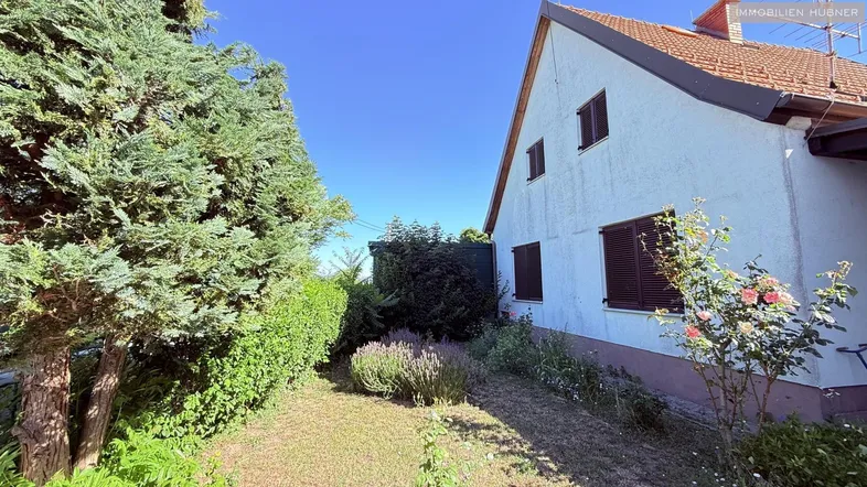 Seitenansicht des Hauses mit gepflegtem Gartenbereich, Bäumen und Sträuchern unter blauem Himmel.