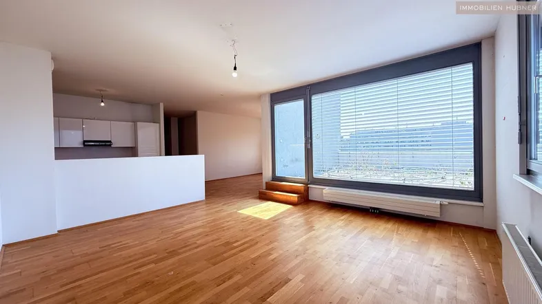 Helles Wohnzimmer mit Holzboden und großem Fenster, angrenzend an eine moderne Küche.