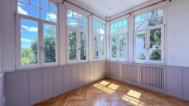 Helles Zimmer mit Fischgrätparkett und vielen Fenstern, die viel Tageslicht hereinlassen.