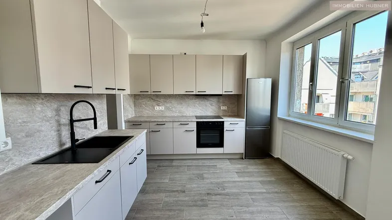 Moderne Küche mit hellgrauen Schränken, schwarzer Spüle, integriertem Backofen und Kühlschrank, sowie einem Fenster.