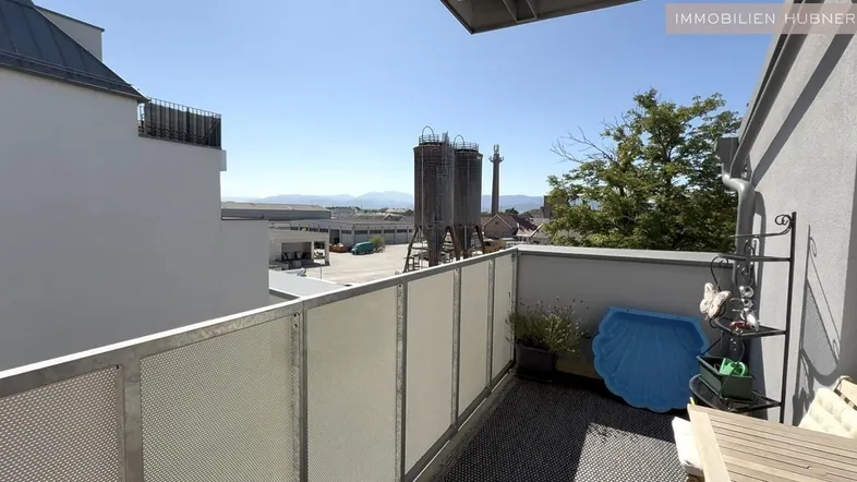 Balkon mit Holztisch und Blick auf Industriegebäude sowie ferne Berge.