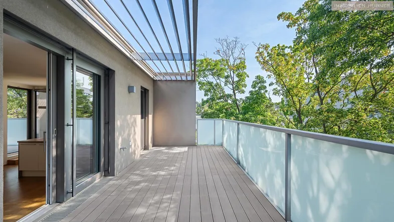 Große Terrasse mit Holzboden und Glasgeländer, umgeben von grünen Bäumen.