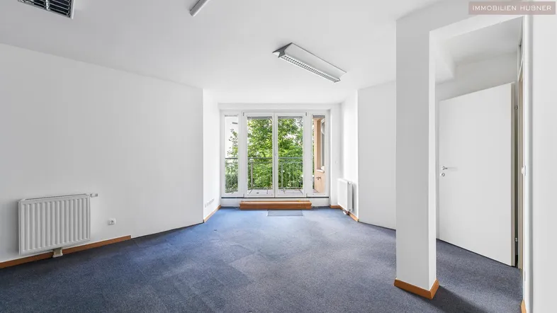 Heller Büroraum mit blauem Teppichboden und Zugang zu einem kleinen Balkon mit Blick ins Grüne.
