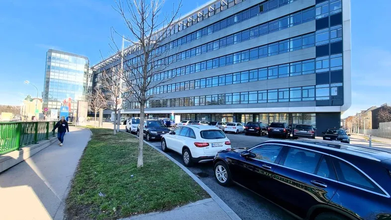 Modernes Bürogebäude mit Glasfassade und Parkplätzen entlang der Straße unter blauem Himmel.