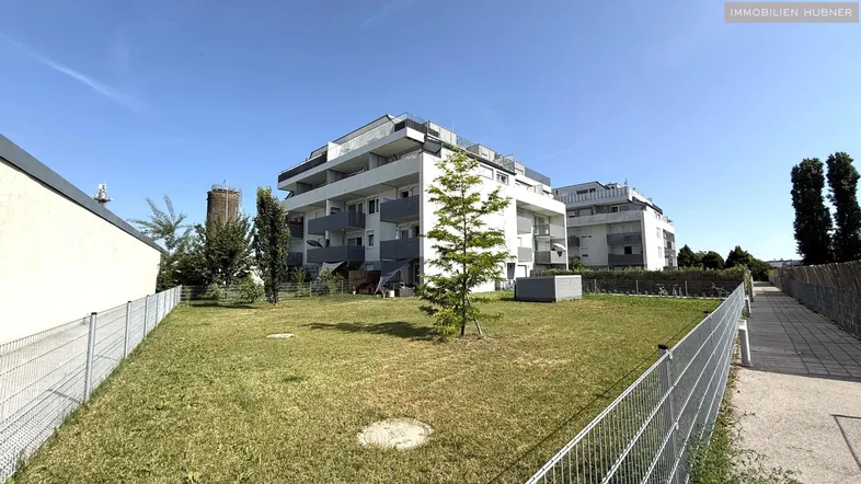 Modernes Mehrfamilienhaus mit Balkonen und einer Grünfläche im Vordergrund unter blauem Himmel.