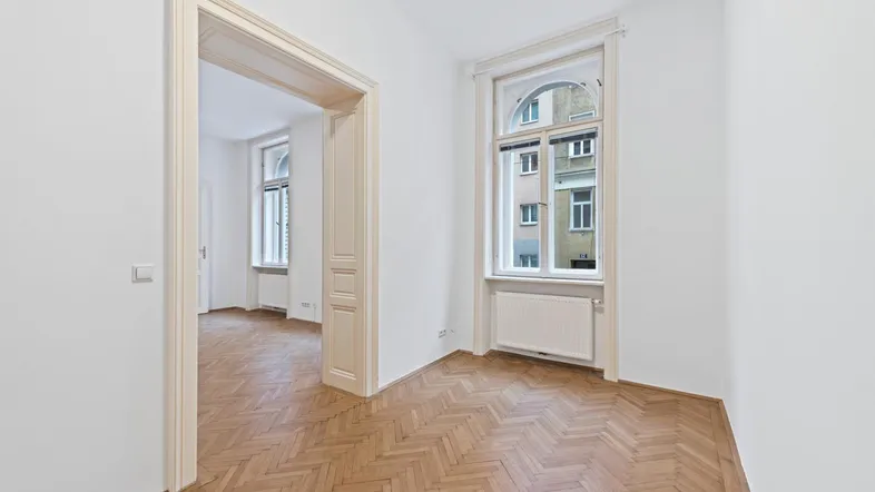 Helles Zimmer mit Fischgrätparkett, hohen Decken und einem großen Fenster mit Blick auf die Straße.