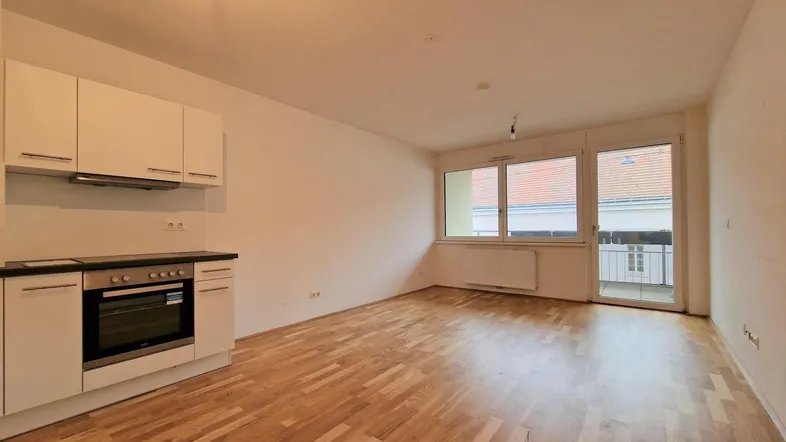 Helles Zimmer mit integrierter Küchenzeile, Parkettboden und Zugang zum Balkon.