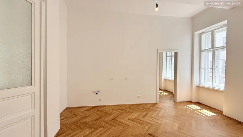 Helles Zimmer mit Fischgrätparkett und hohen Fenstern, bereit für individuelle Gestaltung und Nutzung.