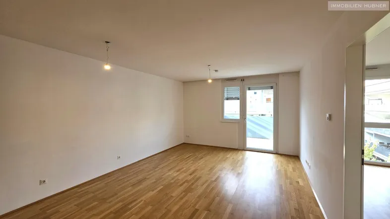 Helles, leeres Wohnzimmer mit Parkettboden und Zugang zu einem Balkon oder einer Terrasse.
