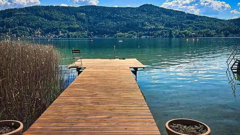 Langer Holzsteg führt in den Wörthersee mit malerischem Bergpanorama unter blauem Himmel.