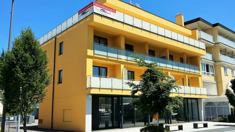 Modernes gelbes Gebäude mit mehreren Balkonen und Geschäften im Erdgeschoss unter blauem Himmel.